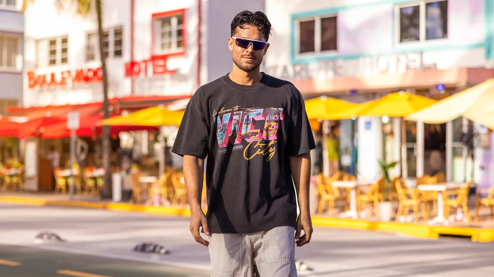 A man in sunglasses on Ocean Drive wearing the black Court Culture x Duvin 'Vice City Postcard' tee with a retro 'Welcome to Vice City' postcard print
